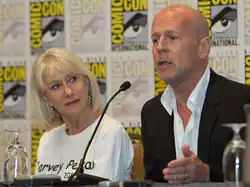 Bruce Willis speaks at San Diego Comic-Con. Actress Helen Mirren is seated to his right wearing a white shirt with the name Harvey Pekar