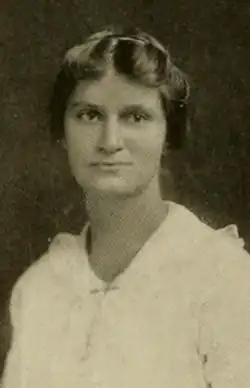 A young white woman wearing a white dress; her hair is parted center and dressed back from her face