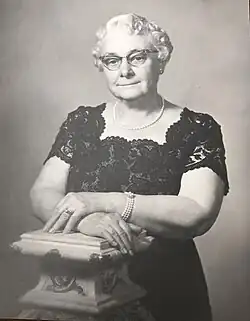 Formal waist-up black-and-white photograph of a white woman in a black lace dress, wearing pearls, her white hair curled, wearing cat-eye glasses. She is leaning on a faux marble Grecian-style column.