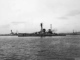 Side view of a large warship with a prominent tripod mast above the superstructure at anchor. Four gun turrets and two funnels are visible.