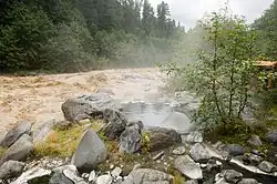Steaming pool of water surrounded by a group of rocks next to a muddy river.