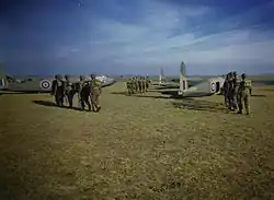 Three groups of six men wearing helmets and backpacks walk across a grass field towards waiting aircraft