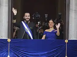 Nayib Bukele and Gabriela Rodríguez de Bukele standing at a balcony and waving to a crowd