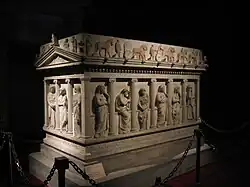 Three quarters view of a decorated marble sarcophagus in the shape of a Greek temple.