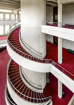 Top of circular staircase ascending anti-clockwise. Made from cream-coloured concrete, floor covered with red carpet.