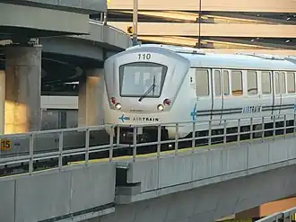 A driverless train on the AirTrain JFK, traveling on a concrete viaduct. There are concrete ramps and buildings behind it.