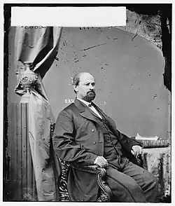 A man with dark hair, a mustache, and a long beard wearing a black jacket and vest and white shirt, sitting in a chair and facing right