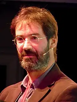 blurry, close-up image of James Oswald wearing a brown jacket, looking just right of camera, with headset microphone