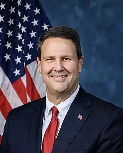 Official House portrait of Crank smiling in front of the U.S. flag, wearing a black suit with American flag lapel pin, white shirt, and red tie.