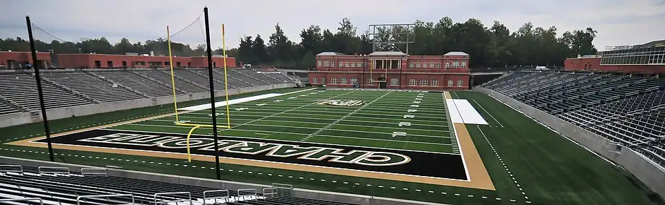 A wide photograph of an empty stadium. Stands are on the left, a brick building behind an American football playing surface is near the center, and home side stands with a press box are on the right.
