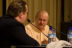 A middle-aged male in a white button-down shirt sits behind a microphone at a convention.