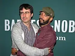 Photo of Oliver, wearing a grey hoodie, embracing Cenac, who is wearing a red hoodie. Both are standing in front of a press backdrop labeled "Barnes & Noble".