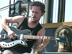 Butler is sitting strumming the strings with his right hand while his left hand is on the fretboard. His hair is relatively short and he is bearded. A microphone is above and to his right with other stage equipment behind his right shoulder. To his left is an oriental statue.