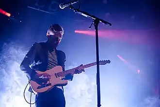 Johnny Blake plays his guitar while standing in front of a microphone. A bright blue light shines on Johnny, and other spotlights in the background aim elsewhere