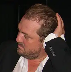 close-up of Jonas Berggren from profile, wearing a white shirt and black blazer, supporting his head with left hand