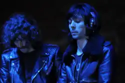 Two men, dressed in black, under a blue light