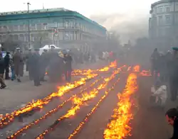 A number of candles are arranged in a cross shape in a roadway, while a crowd of people look on.