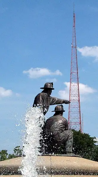 A tall, red lattice tower against a blue sky at day with a fountain and sculpture in the foreground.
