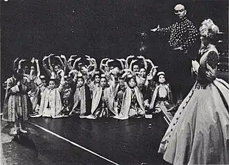 Black and white photo of a theatrical scene: a middle-aged man with a shaved head and imposing presence has his right arm extended to introduce a group of children in Asian dress to a woman in a crinoline dress and bonnet in the foreground at right, who is partially turned upstage. The children are mostly kneeling and have their arms raised in greeting; one child (probably Crown Prince Chulalongkorn) stands and bows.