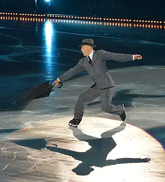 A man wearing a gray suit and fedora and holding an umbrella skates on ice