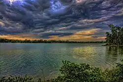 Gray storm clouds roll over green trees and plants that rim a choppy gray lake