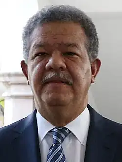 A portrait shot of a middle-aged man smiling somewhat and looking straight ahead. He has light brown skin, slightly African facial features, curly dark hair. He is mustachioed and wears a suit and tie.