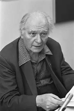 A black-and-white photo of an elderly, balding man with swept-back hair, wearing a suit; he faces the camera.