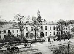 A black and white photograph, showing a stately-looking two storey building with white walls, extending out of shot to the left and right, with an arched cart entrance at the centre. A modest clocktower rises above the entrance, and the building is surrounded by neat shrubbery and iron railings. A wide street crosses left-right outside of the fence, with a handful of horse-drawn carts and pedestrians in 19th century clothing.