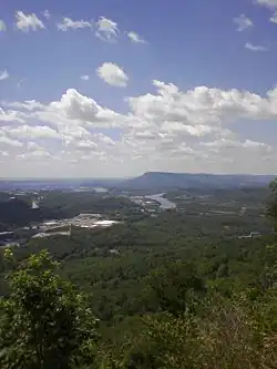 View from high up on a mountain with buildings alongside river bends and indistinct forested land in middle distance stretching towards hazy indistinct mountain across the horizon in distance on a bright day with blue and white dappled sky