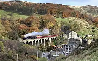 A steam train running across a stone viaduct, with steep hills behind, and mill buildings in the foreground