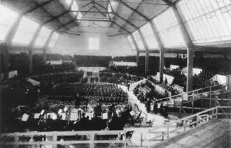 Darkened interior of a large hall with two rows of high windows along each side. It is possible to discern a seated orchestra in the foreground, with mass choirs in the background.