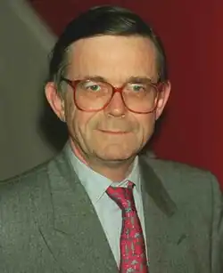 cleanly-shaven caucasian male aged in his 50s with greased-back hair, wearing a light grey suit, red tie, and large red spectacles