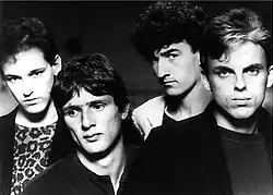 Head and shoulders shot of four men. Man at left has dark hair, face slight tilted to his right, but looking forward, he wears a dark jacket over a print tee-shirt. Second man is the shortest, also dark haired, his head is tilted further with eyes looking to his right, he wears a dark tee-shirt. Third man is the tallest, also dark haired, with his face partly in left profile, his head is also titled, his eyes are direct to the viewer, he wears a light coloured jacket with a dark tee-shirt. The last man is facing forward, he is the second tallest, his hair is lighter with fair coloured streaks at front, his jacket is dark over a less dark tee-shirt.