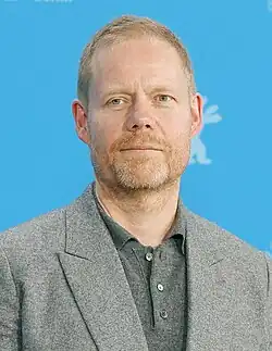 A middle-aged man with reddish hair and light beard wearing a gray polo under a gray jacket, photographed against a blue background