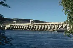 A concrete hydroelectric dam's spillway on the downriver side. Water is in the foreground and power lines run overhead.