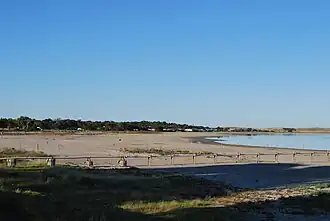 The picture shows Lake Albert from its shore. In the foreground is a sandy shore and a pipeline leading towards the lake. The lake is in the right of the photo. In the background there are trees on the left and brown hills on the right.