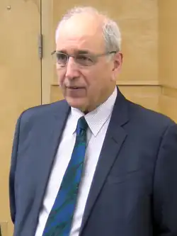 Half-length photoportrait of Lynk standing. He is wearing a dark blue suit with a blue-green tie.