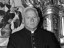 A balding man in priest garb looks absentmindedly near the camera's focal point