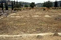Image of the remains of the first oikos taken in 1991. The west wall is in the foreground and the limestone walls are very short and damaged. Only the rectangular walls remain around the soil and grass, there are also hills and trees in the background.