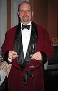 A white man with a goatee is wearing a maroon-colored jacket that ties around the middle with black ribbon. The lapels are wide and black. The jacket is open to the waist, showing a white, high collared shirt beneath with a black bowtie. The man is holding two champagne glasses.