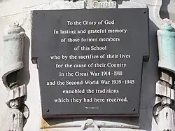 Affixed to the weathered, discoloured white stone war memorial is a black plaque with white text in Times New Roman font. The text, which is centred and has generous line spacing, reads: "To the Glory of God / In lasting and grateful memory / of those former members / of this School / who by the sacrifice of their lives / for the cause of their Country / in the Great War 1914 – 1918 / and the Second World War 1939 – 1945 / ennobled the traditions / which they had here received."