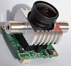 Image of a machine attached to a circuit board. The machine has a round black component mounted onto a metal block.