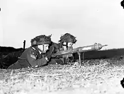 Two men in military uniform lie on a grassy bank, handling a tube-shaped weapon