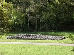 Elevated view of the cairn in the middle distance, from its side, with deciduous trees in leaf to its rear. To its front passes a wide asphalt path, dissecting flat ground of short grass. The tumulus' trapezium shape is evident, its boulders retained by a short wall, missing at the very front, left, where the rubble has tumbled out.