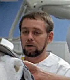 A man with short brown hair, beard and mustache wearing a white top looks to the left and down; he appears to be in a hospital room