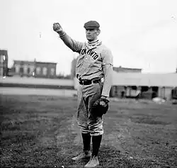 A black-and-white photograph of a man wearing an old-style baseball uniform and a newsie cap holding a baseball in his outstretched hand