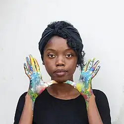 Self-portrait of Sardoine Mia, a young black woman with dark curly hair wearing a black headcloth and shirt. She is standing in front of a white wall, posing with her hands covered in many types of paint.