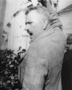 An image showing an older middle-aged man with grey hair and moustache, raising his hand and giving a passionate speech.