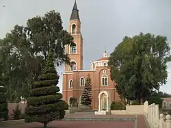 Orange coloured church with steeple visible between trees and a courtyard in the foreground