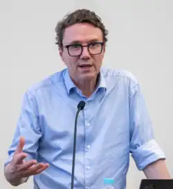 Jens Nielsen wearing a light blue shirt, speaking into a microphone at a podium, gesticulating with right hand